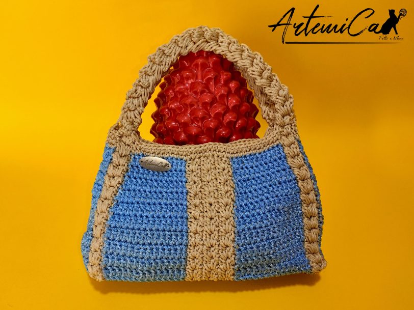 1 L'immagine mostra una borsa azzurra all'uncinetto con strisce verticali gialle, dotata di un manico coordinato e una chiusura magnetica. Sogno Mediterraneo: il fascino del mare nella tua mano.