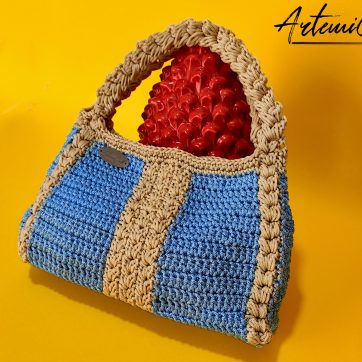 L'immagine mostra una borsa azzurra all'uncinetto con strisce verticali gialle, dotata di un manico coordinato e una chiusura magnetica. Sogno Mediterraneo: il fascino del mare nella tua mano.