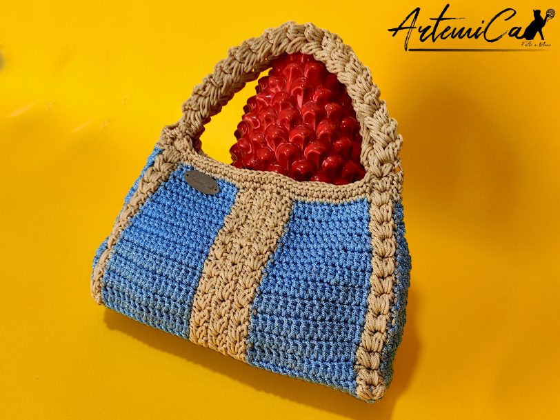 2 L'immagine mostra una borsa azzurra all'uncinetto con strisce verticali gialle, dotata di un manico coordinato e una chiusura magnetica. Sogno Mediterraneo: il fascino del mare nella tua mano.