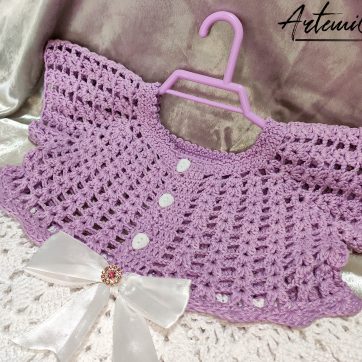 Il Vestitino handmade per bambina su Artemicat.com è ritratto in tutta la sua bellezza. La giacchetta a mezza manica in lilla chiaro si abbina splendidamente alla gonnellina bianca, mentre il fiocco decorativo al centro aggiunge un tocco di eleganza. I dettagli artigianali, come i bottoni dorati e il lavoro all'uncinetto, sono chiaramente visibili, evidenziando la qualità e l'amore dedicato alla creazione di questo incantevole capo per bambini.