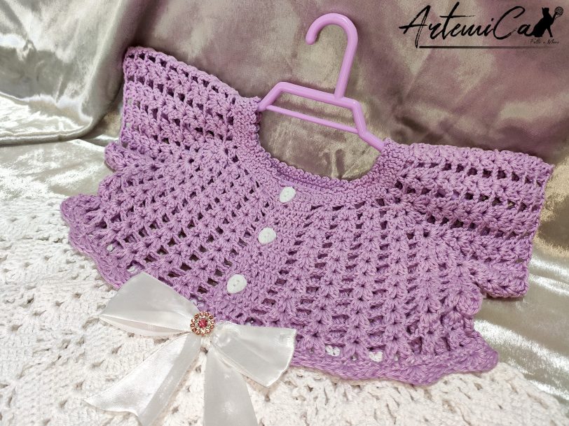 2 Il Vestitino handmade per bambina su Artemicat.com è ritratto in tutta la sua bellezza. La giacchetta a mezza manica in lilla chiaro si abbina splendidamente alla gonnellina bianca, mentre il fiocco decorativo al centro aggiunge un tocco di eleganza. I dettagli artigianali, come i bottoni dorati e il lavoro all'uncinetto, sono chiaramente visibili, evidenziando la qualità e l'amore dedicato alla creazione di questo incantevole capo per bambini.