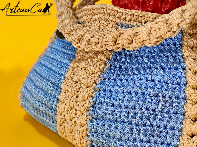 4 L'immagine mostra una borsa azzurra all'uncinetto con strisce verticali gialle, dotata di un manico coordinato e una chiusura magnetica. Sogno Mediterraneo: il fascino del mare nella tua mano.